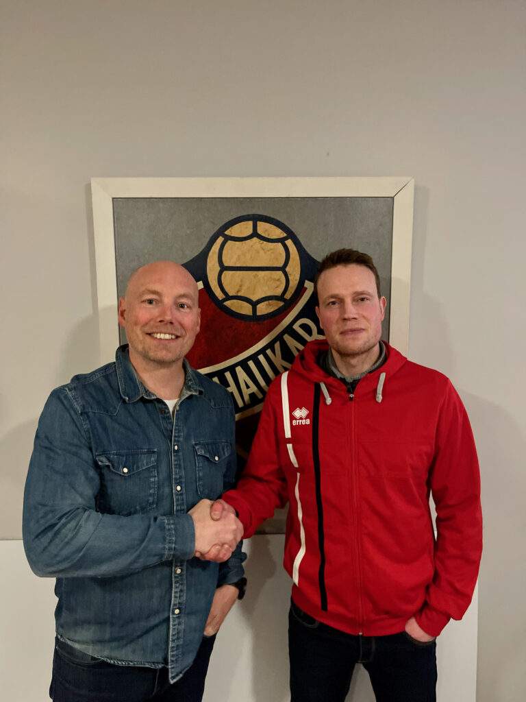 Tveir karlmenn takast í hendur fyrir framan merki Hauka. 